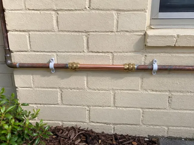 Repaired copper water pipe on a painted brick exterior wall with brackets and a window in Peakhurst