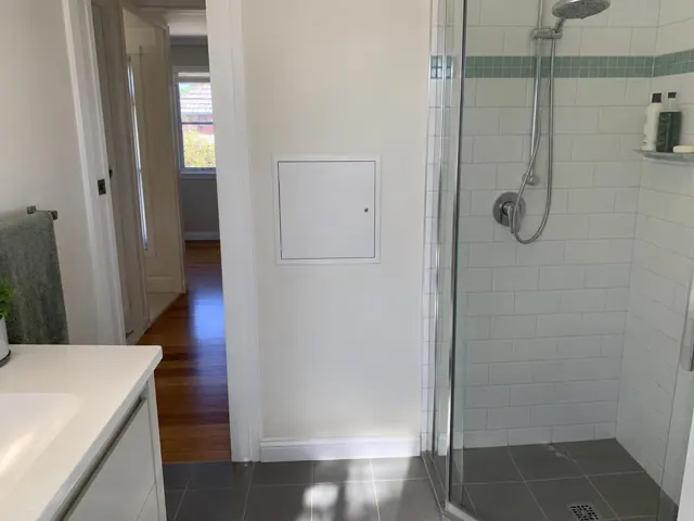 Bathroom wall with a small access panel beside a glass shower and tiled floor in Hurstville